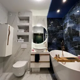 Elegancka łazienka z marmurowymi płytkami, okrągłym lustrem z podświetleniem LED, wolnostojącą wanną z drewnianą półką i złotą armaturą.