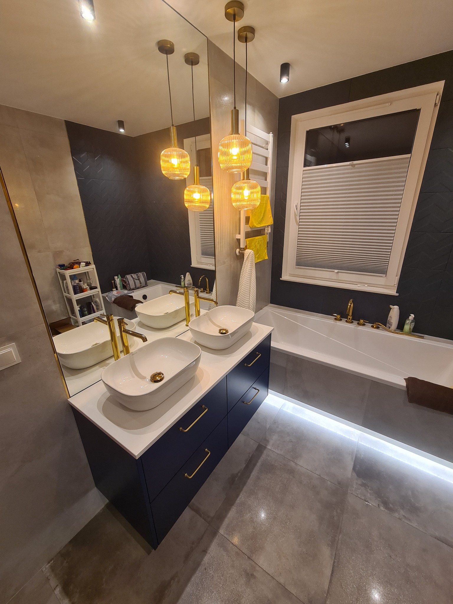 Nowoczesna łazienka z dwoma umywalkami nablatowymi na granatowej szafce, złotymi bateriami i oświetleniem, szare płytki podłogowe z podświetleniem LED, wanna pod oknem z żaluzjami plisowanymi.