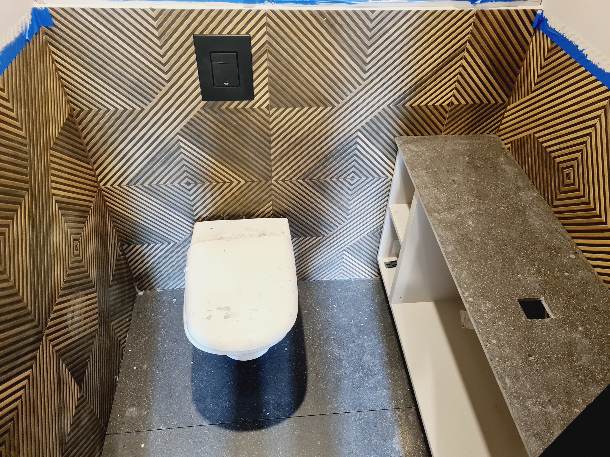 Nowoczesna toaleta z geometryczną okładziną ścienną w odcieniach brązu i beżu, czarny przycisk spłuczki, szara podłoga z płytek, fragment blatu z otworem, pomieszczenie w trakcie wykańczania...