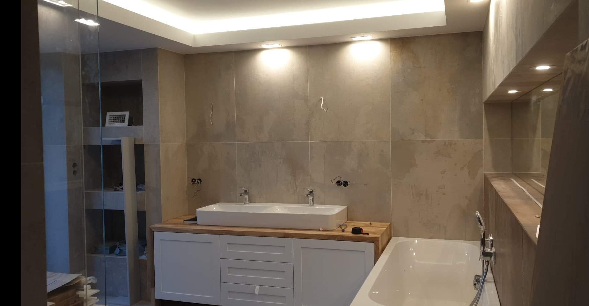 Nowoczesna łazienka z beżowymi płytkami imitującymi kamień, podwójną umywalką nablatową na drewnianym blacie z białymi szafkami, kabiną prysznicową ze szklaną ścianą i wanną z chromowaną baterią.