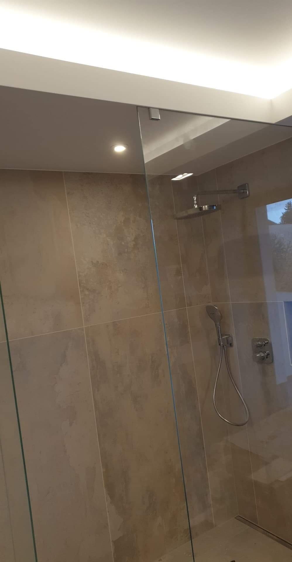 Beżowe płytki w kabinie prysznicowej z deszczownicą i słuchawką, widok przez szklaną ścianę, oświetlenie sufitowe LED.