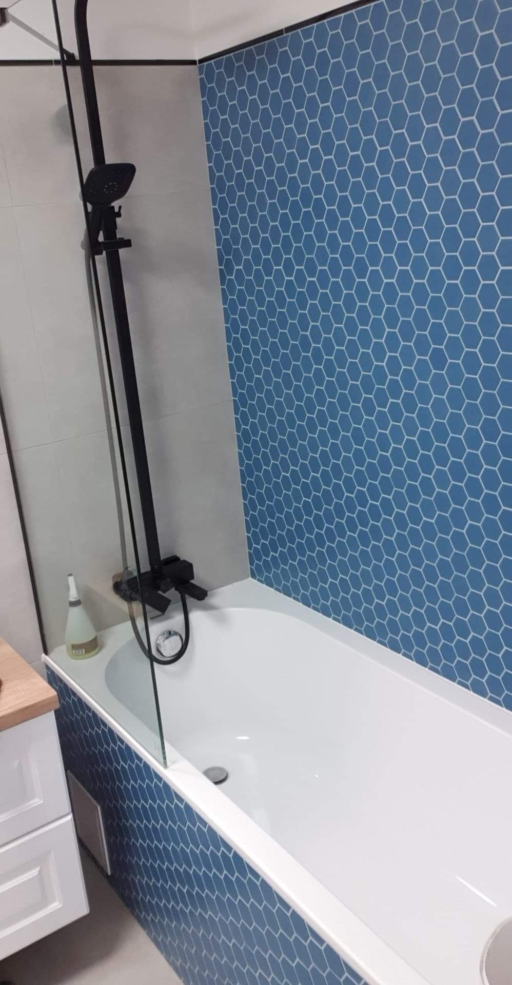 Nowoczesna łazienka z wanną obłożoną niebieskimi heksagonalnymi płytkami, czarna armatura prysznicowa, szklany parawan nawannowy.