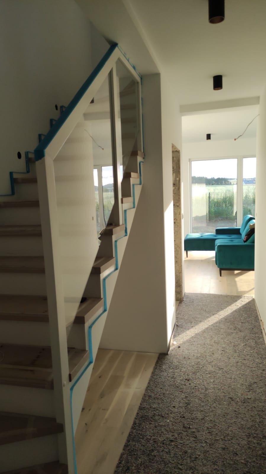 Wnętrze domu w trakcie wykańczania: drewniane schody z białymi balustradami zabezpieczone niebieską folią, widok na salon z turkusową sofą w tle, podłoga zabezpieczona materiałem budowlanym.