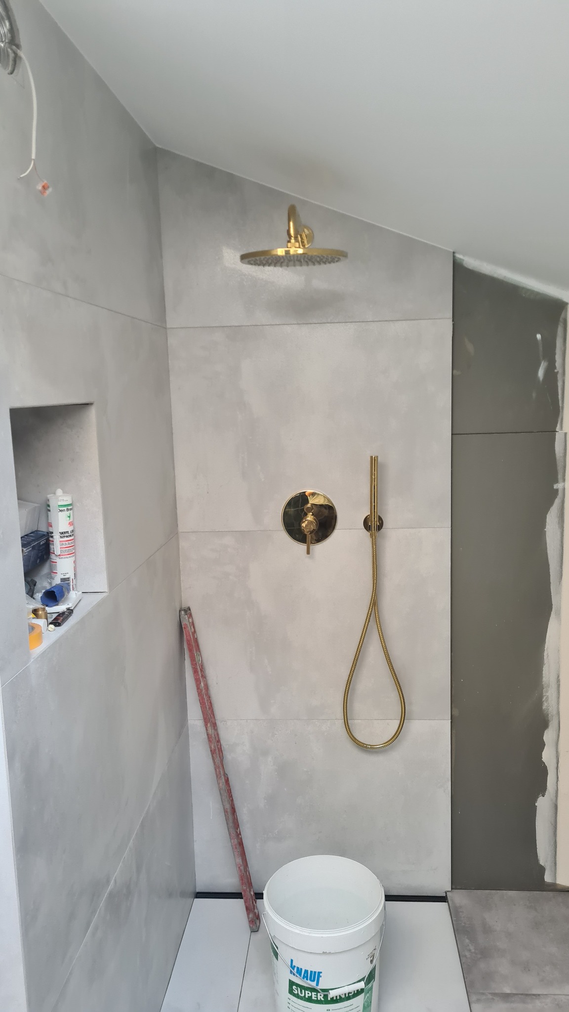 Szare płytki w kabinie prysznicowej z złotą armaturą, widoczny kubeł z masą szpachlową Knauf i poziomica oparta o ścianę.
