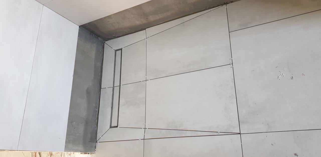 Układanie szarych, matowych płytek podłogowych w łazience z odpływem liniowym; widoczne krzyżyki dystansowe.