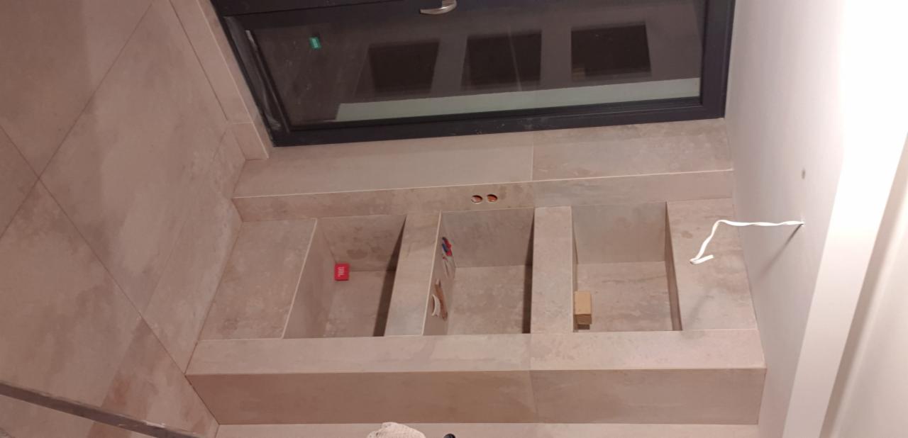 Niedokończona zabudowa umywalki z beżowych płytek ceramicznych, widoczne wystające kable i otwór okienny w suficie.