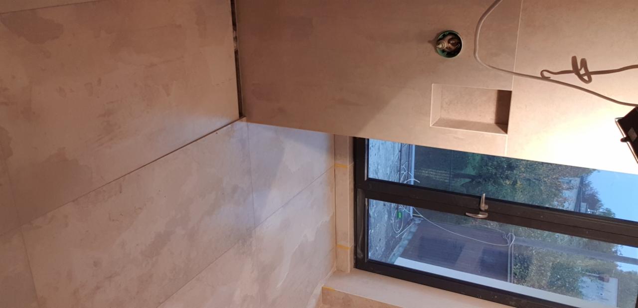 Ściana i podłoga w trakcie układania płytek w łazience, widoczna wnęka w ścianie, niedokończone fugowanie, okno z widokiem na zewnątrz, wystające kable elektryczne.