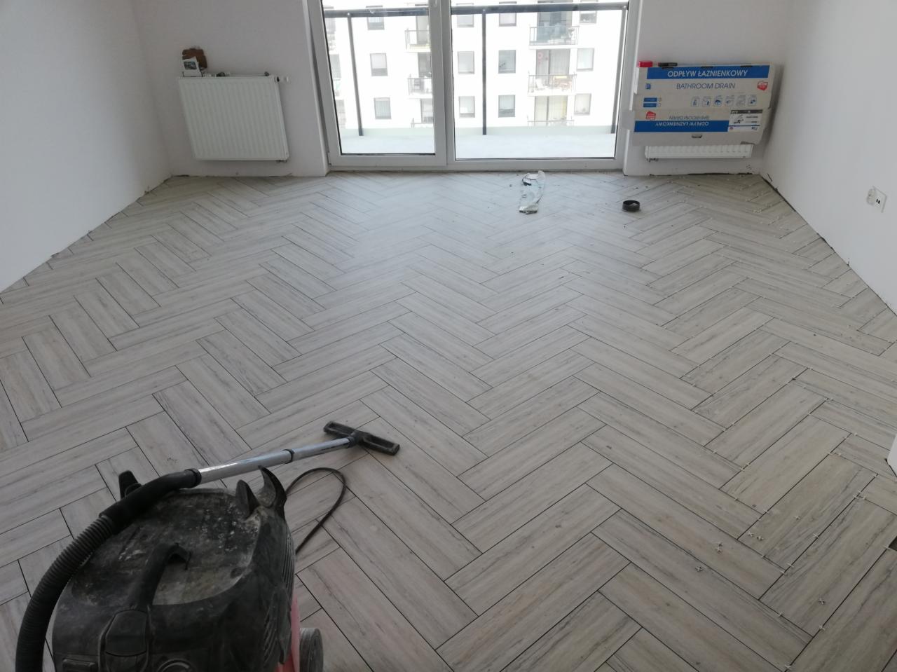 Ułożona podłoga z płytek imitujących drewno w jodełkę w jasnym pomieszczeniu z dużym oknem balkonowym, widoczny odkurzacz przemysłowy i karton z odpływem łazienkowym.