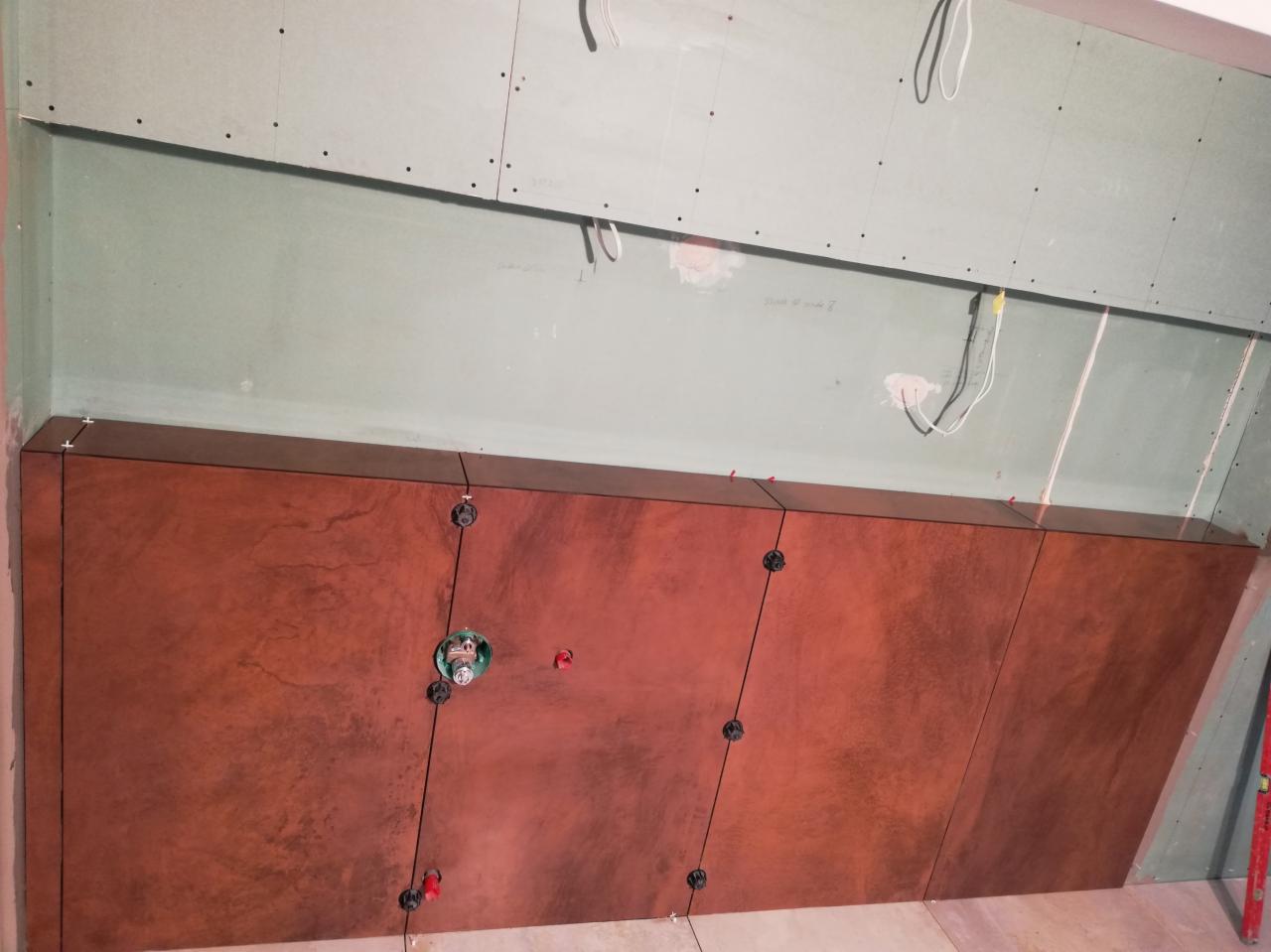 Montaż brązowych płytek ściennych z marmurkowym wzorem, widoczne krzyżyki dystansowe i otwór na instalację hydrauliczną, surowa ściana z płyt kartonowo-gipsowych w tle.