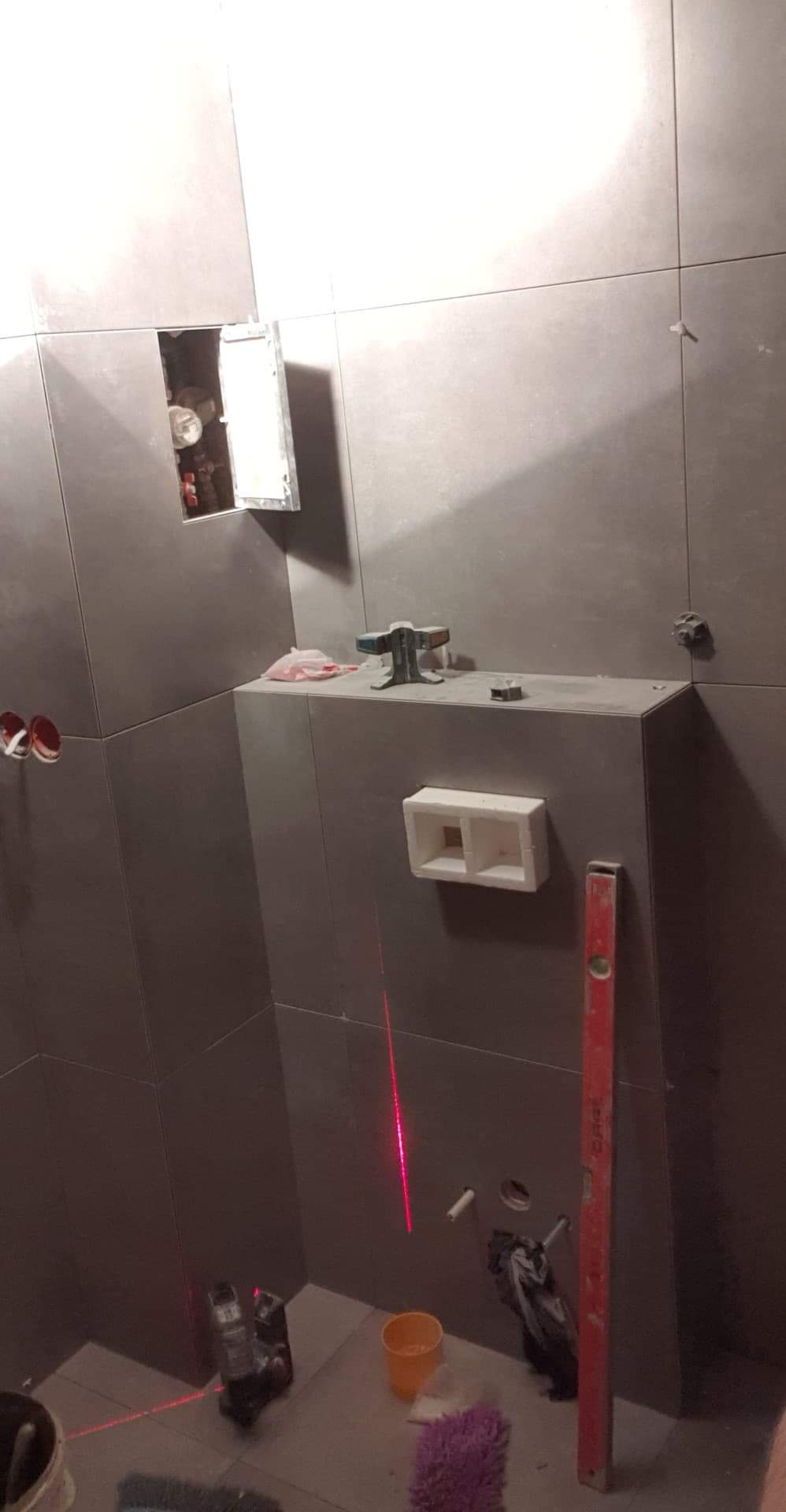 Montaż szarej glazury w łazience, widoczny laserowy niwelator, poziomica, otwór rewizyjny i puszka podtynkowa.