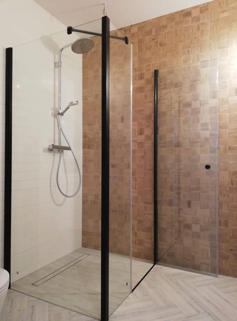 Nowoczesna kabina prysznicowa z czarnymi profilami, deszczownicą i szklanymi ścianami, na tle ściany wyłożonej mozaiką z drewna w odcieniach brązu i beżu. Podłoga wyłożona płytkami imitującymi...