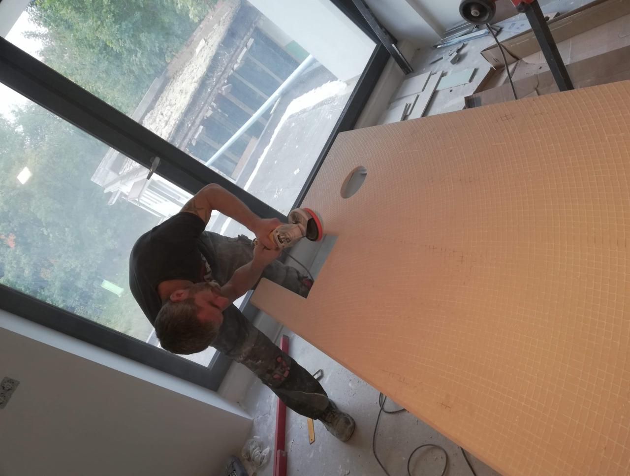 Pracownik szlifuje płytę z mozaiki ceramicznej za pomocą szlifierki kątowej, przygotowując otwór na armaturę; widoczne narzędzia i okno z widokiem na zieleń.
