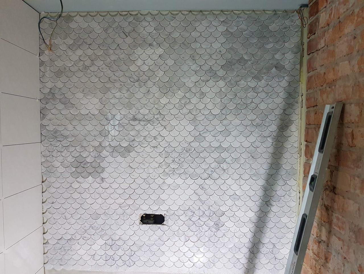 Ściana w łazience wyłożona mozaiką w kształcie rybiej łuski, częściowo wykończona, z widoczną puszką elektryczną i poziomnicą opartą o surową cegłę.