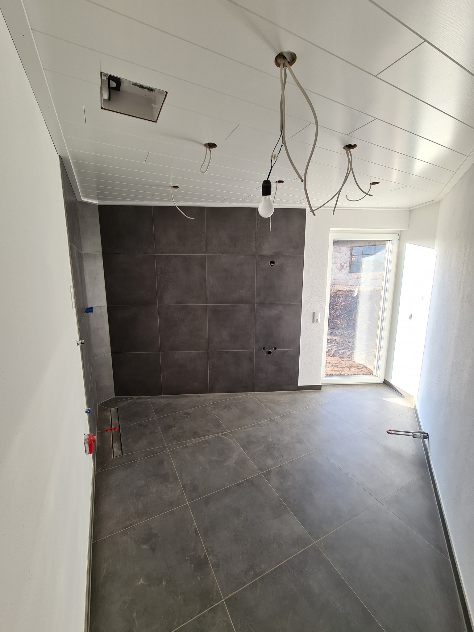 Wnętrze w trakcie remontu: szare płytki na ścianie i podłodze, biały sufit z wystającymi kablami i otworem instalacyjnym, widoczne narzędzia przy ścianie, okno z widokiem na zewnątrz.