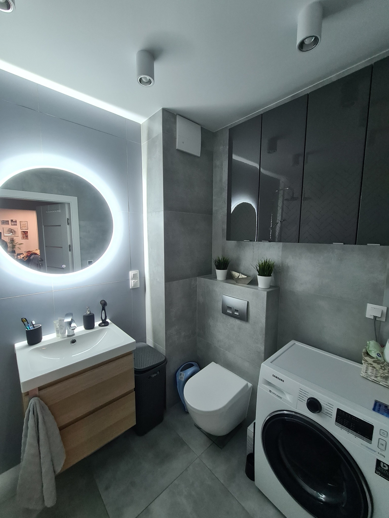 Nowoczesna łazienka w szarościach z okrągłym lustrem podświetlanym LED, drewnianą szafką pod umywalką, wiszącą toaletą i pralką Samsung.