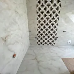 Wnętrze łazienki w trakcie wykańczania: marmurowe płytki na ścianach i podłodze z charakterystycznym panelem dekoracyjnym z motywem kół w odcieniach brązu i beżu. Widoczne otwory na armaturę...