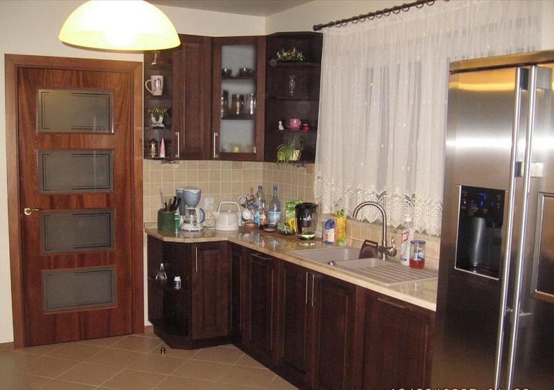 Kadr z kuchni z ciemnymi, drewnianymi szafkami, jasnym blatem i dużym, srebrnym lodówką; widoczna również drewniana stolarka drzwiowa z trzema matowymi szybami.