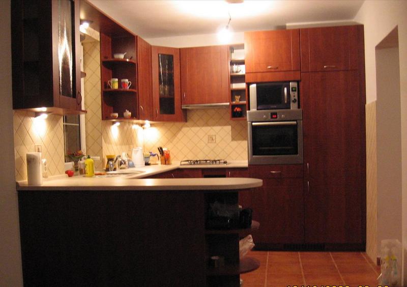 Kuchnia z zabudową w odcieniach brązu, z oświetleniem punktowym pod szafkami wiszącymi i blatem roboczym, wyposażona w piekarnik i mikrofalówkę w zabudowie oraz półwysep z barkiem.