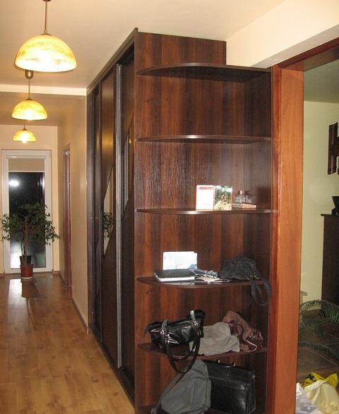 Wnętrze przedpokoju z dużą szafą przesuwną w ciemnym drewnie i otwartymi półkami narożnymi, oświetlone trzema lampami wiszącymi, widok na jasne drzwi balkonowe i roślinę doniczkową.