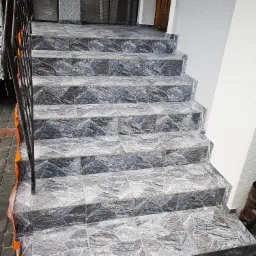 Schody zewnętrzne wyłożone szarą, wzorzystą płytką ceramiczną, z metalową balustradą po lewej stronie i fragmentem elewacji w tle.