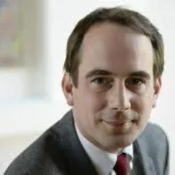 Portret mężczyzny w garniturze i czerwonym krawacie, uśmiechającego się do kamery, z obrazem w tle.