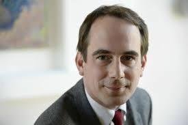 Portret mężczyzny w garniturze i czerwonym krawacie, uśmiechającego się do kamery, z obrazem w tle.