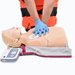 Instruktor w pomarańczowym mundurze demonstruje resuscytację krążeniowo-oddechową na manekinie z podłączonym defibrylatorem, ręce w niebieskich rękawiczkach ułożone na klatce piersiowej.