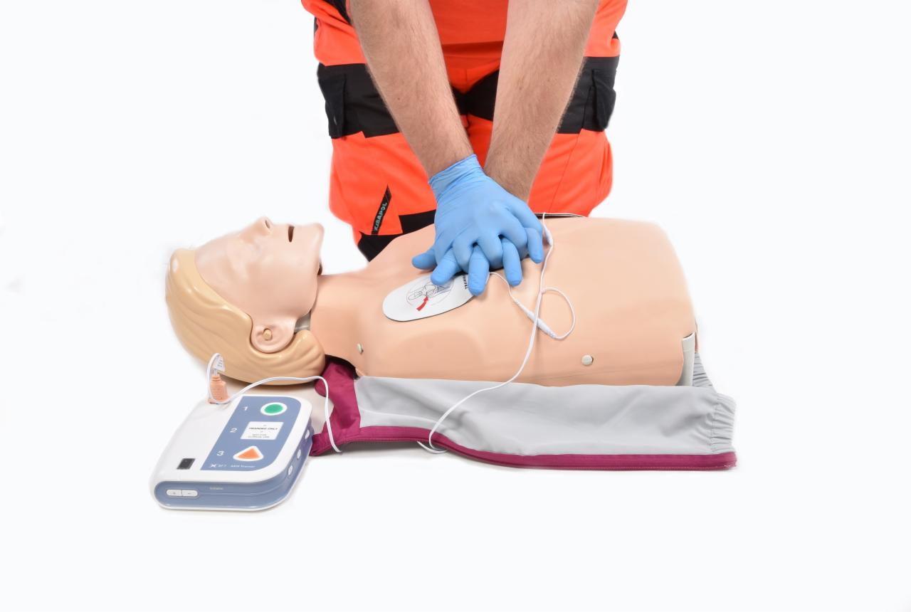 Instruktor w pomarańczowym mundurze demonstruje resuscytację krążeniowo-oddechową na manekinie z podłączonym defibrylatorem, ręce w niebieskich rękawiczkach ułożone na klatce piersiowej.