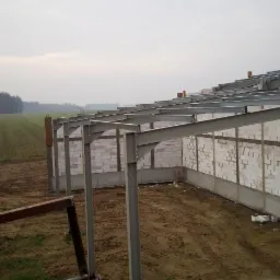 Wznoszenie konstrukcji budynku: ściany z białych bloczków, stalowy szkielet dachu, widok na pole i las w tle, pochmurny dzień.