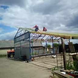 Trzech dekarzy na dachu metalowej konstrukcji w trakcie krycia, widoczny traktor i budynki gospodarcze w tle, pochmurne niebo.