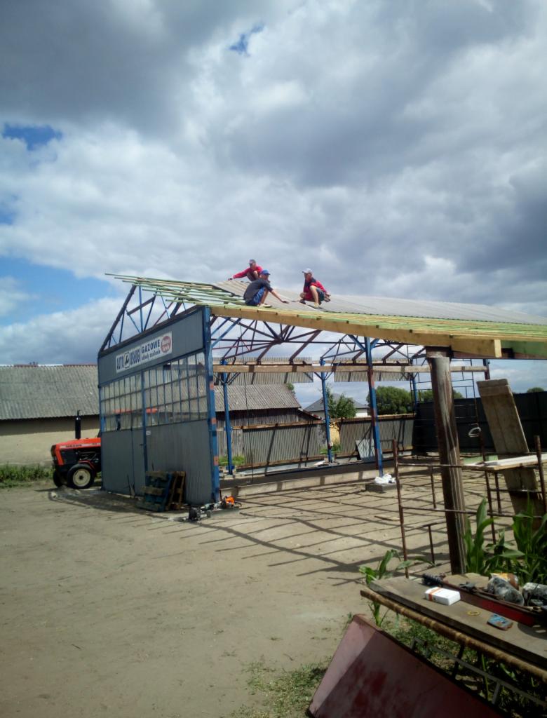 Trzech dekarzy na dachu metalowej konstrukcji w trakcie krycia, widoczny traktor i budynki gospodarcze w tle, pochmurne niebo.
