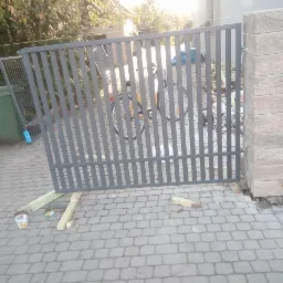 Montaż szarej, metalowej bramy wjazdowej z pionowymi szczebelkami na posesji z kostki brukowej, widoczne tymczasowe podpory z drewna i filar bramy z bloczków betonowych.