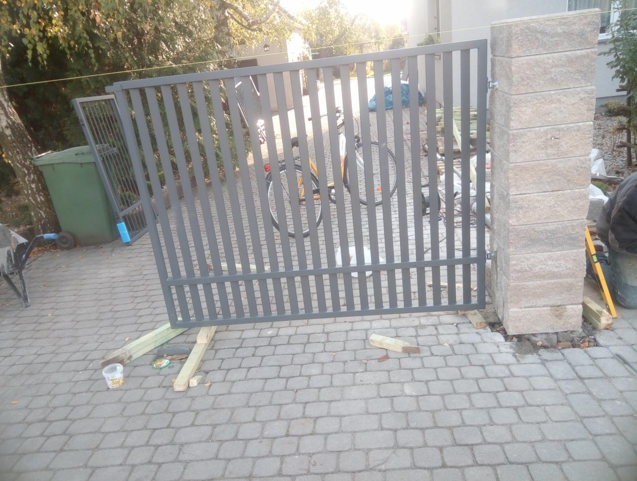 Montaż szarej, metalowej bramy wjazdowej z pionowymi szczebelkami na posesji z kostki brukowej, widoczne tymczasowe podpory z drewna i filar bramy z bloczków betonowych.