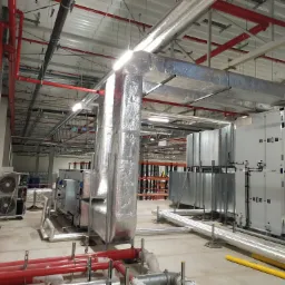 Instalacja wentylacyjna w hali przemysłowej z widocznymi przewodami izolowanymi folią aluminiową, czerwonymi rurami oraz centralą wentylacyjną z dwoma wentylatorami.