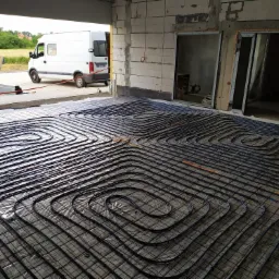 Rozłożona instalacja ogrzewania podłogowego z rur grzewczych na folii, widoczny samochód dostawczy na zewnątrz budynku w stanie surowym.