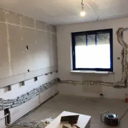 Wnętrze w trakcie remontu: rury instalacyjne prowadzone wzdłuż ścian, częściowo ukryte pod płytami gipsowo-kartonowymi, widoczne narzędzia murarskie na podłodze, okno z roletami.