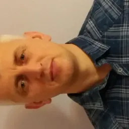 Portret mężczyzny z krótkimi, jasnymi włosami w niebieskiej koszuli w kratę, z delikatnym uśmiechem, spoglądającego w obiektyw na jasnym tle.