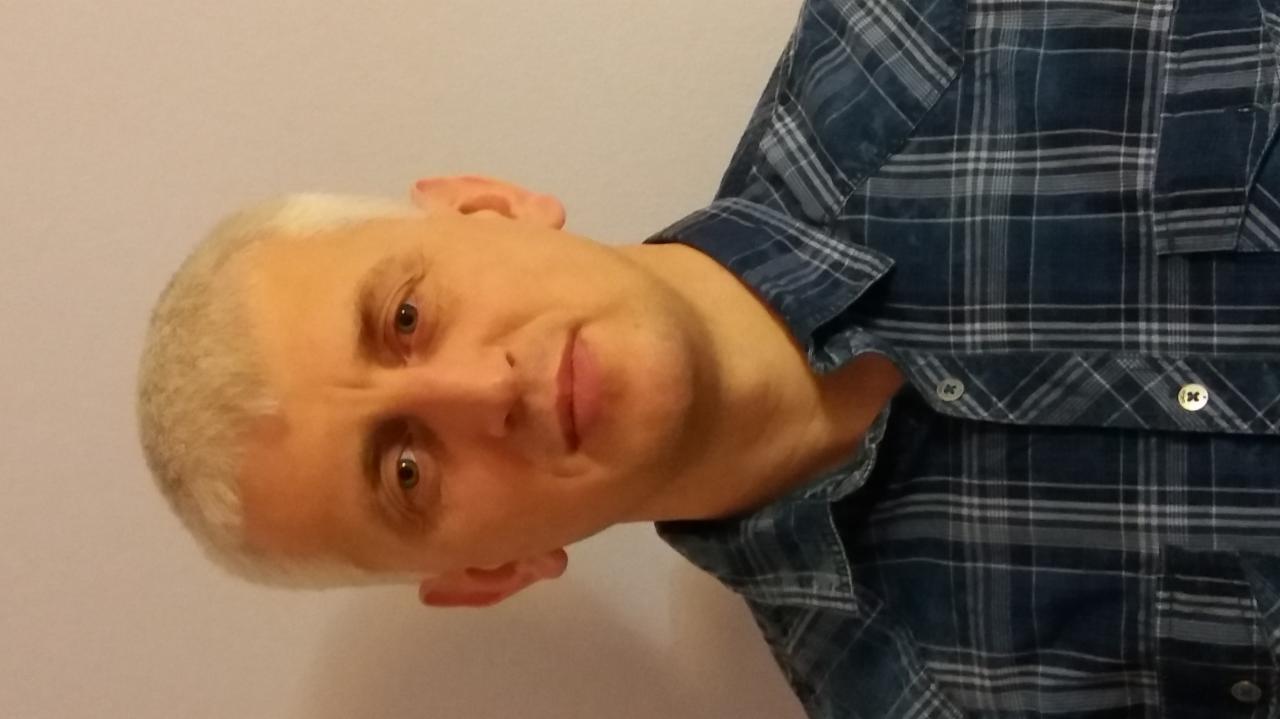 Portret mężczyzny z krótkimi, jasnymi włosami w niebieskiej koszuli w kratę, z delikatnym uśmiechem, spoglądającego w obiektyw na jasnym tle.