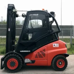 Pomarańczowo-czarny wózek widłowy Linde H50D na zewnątrz, widok z boku, z naklejką informacyjną na karoserii. Na drugim planie siatka zabezpieczająca.