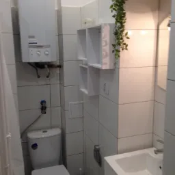 Wnętrze małej łazienki z białymi płytkami, wiszącym piecykiem gazowym Vaillant, toaletą, umywalką i wiszącymi p&oacute;łkami dekorowanymi sztuczną rośliną.