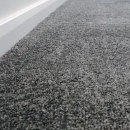 Nowa, szara wykładzina dywanowa z niskim włosiem, przylegająca do białej listwy przypodłogowej w jasnym pomieszczeniu.