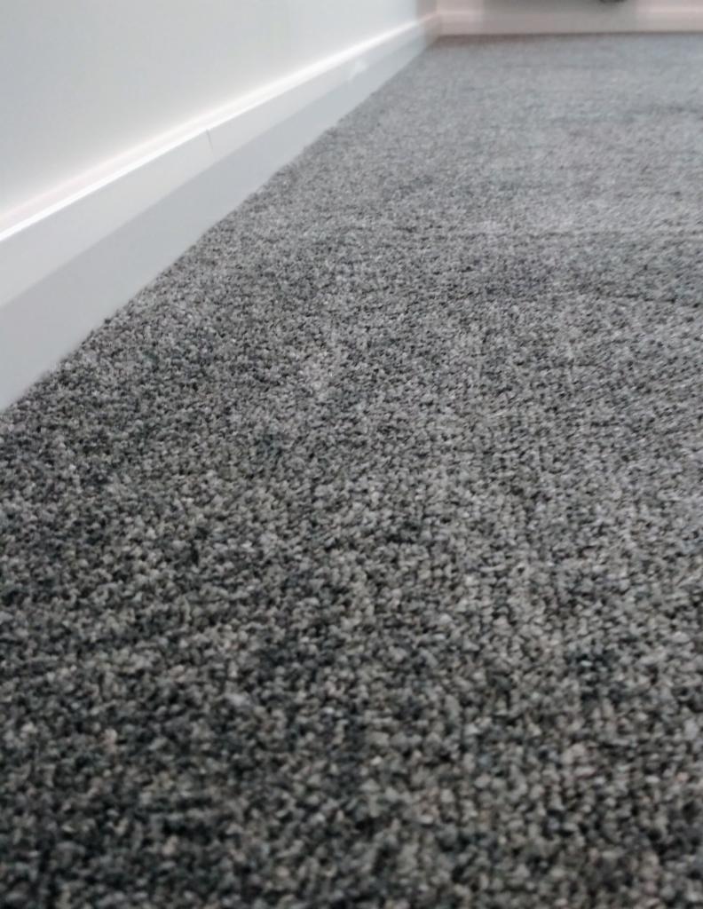 Nowa, szara wykładzina dywanowa z niskim włosiem, przylegająca do białej listwy przypodłogowej w jasnym pomieszczeniu.
