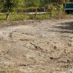 Przygotowany teren pod budowę z widocznymi śladami opon i resztkami gałęzi, na drugim planie żółty samochód ciężarowy z zieloną plandeką.