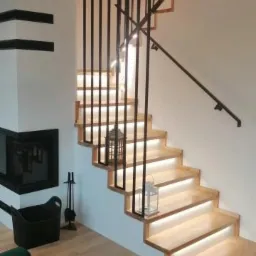 Nowoczesne schody drewniane z podświetleniem LED stopni i czarnymi balustradami, obok kominek z czarną obudową i dekoracyjna latarenka na schodach.