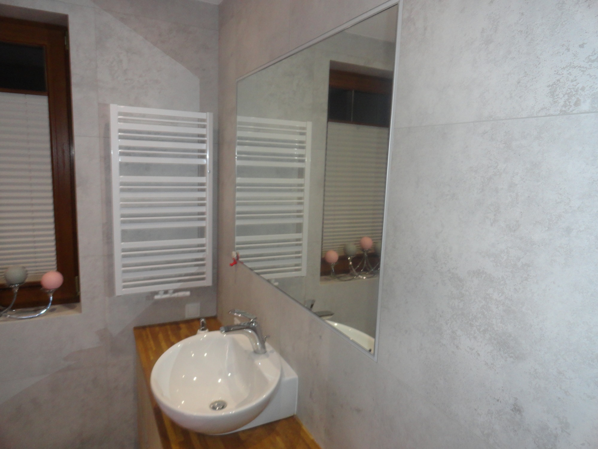 Wykończona łazienka z owalną umywalką nablatową, drewnianym blatem, grzejnikiem drabinkowym i dużym lustrem bez ramy, płytki imitujące beton na ścianach.