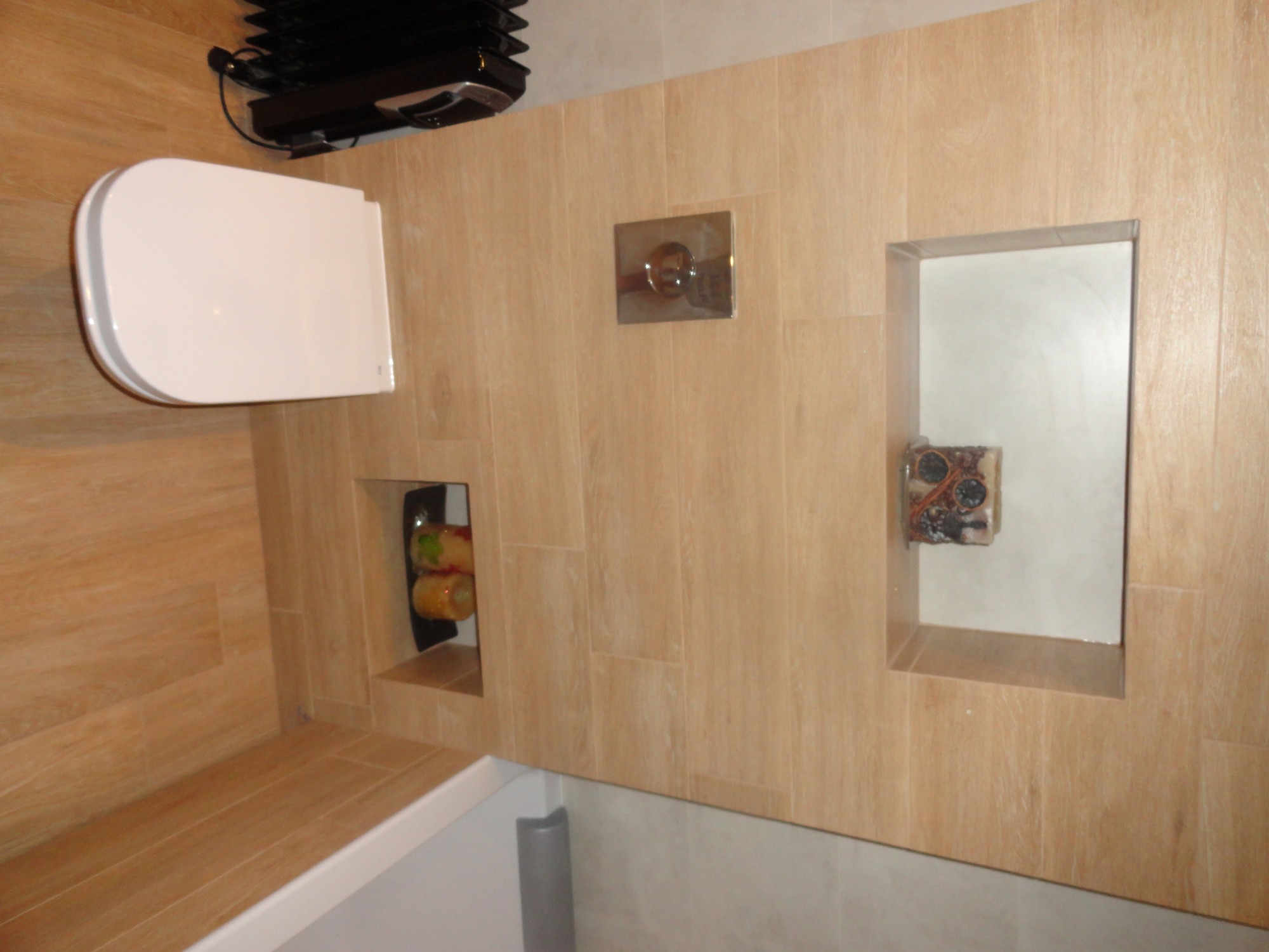 Wykończona łazienka z toaletą podwieszaną, płytkami imitującymi drewno, wnękami dekoracyjnymi z ozdobami i chromowanym przyciskiem spłukiwania.