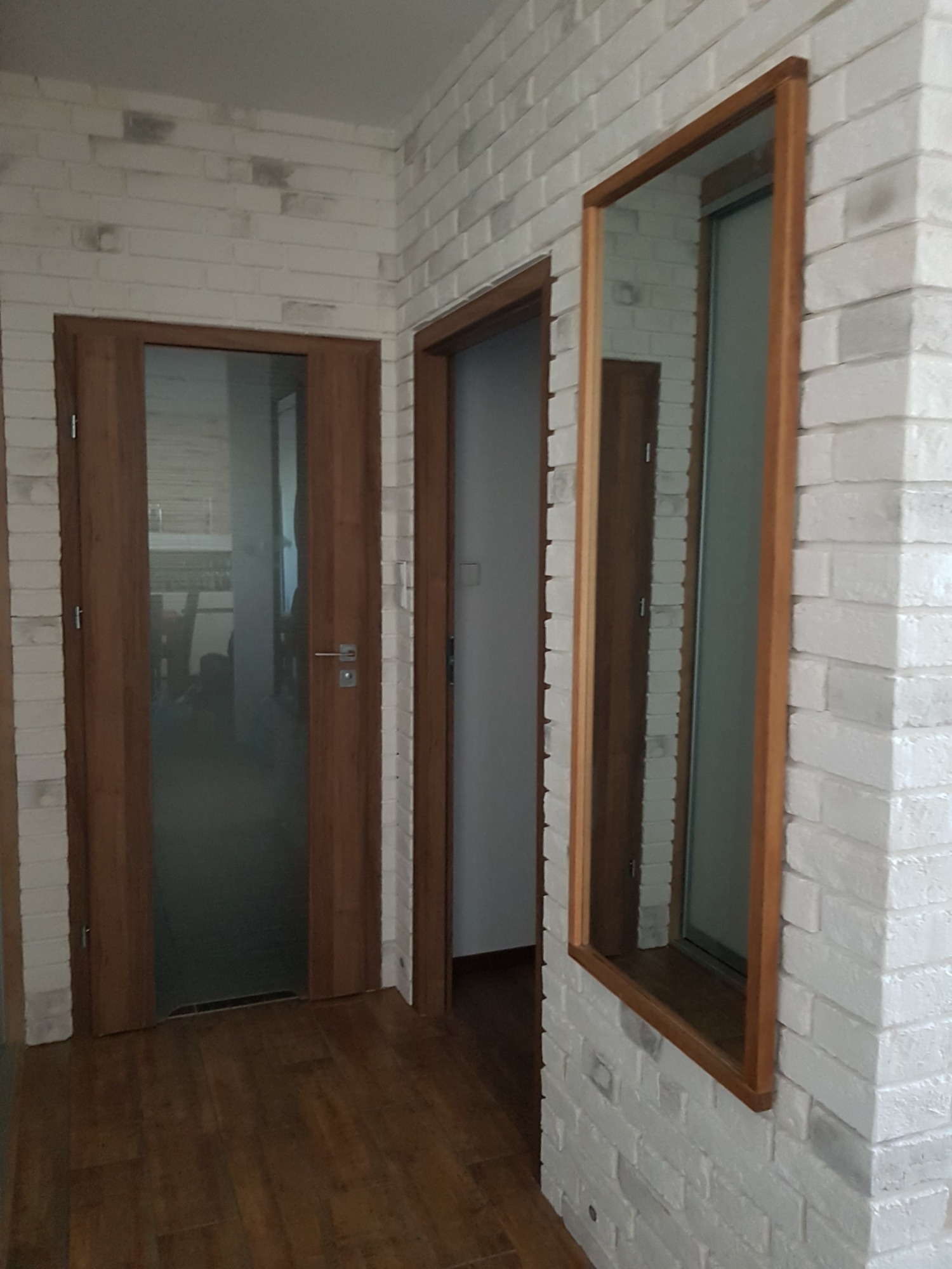 Wnętrze z białej cegły z dwiema drewnianymi ościeżnicami drzwi i lustrem w drewnianej ramie, widoczne drewniane panele podłogowe.