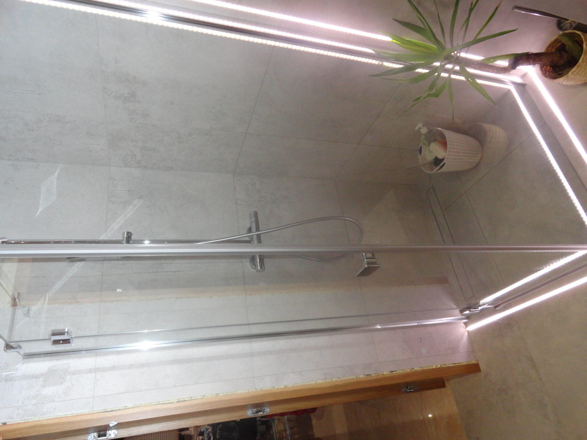 Kabina prysznicowa ze szklanymi drzwiami i chromowanymi okuciami, wykończona jasnoszarą płytką imitującą kamień, z oświetleniem LED wzdłuż sufitu i rośliną doniczkową w rogu.