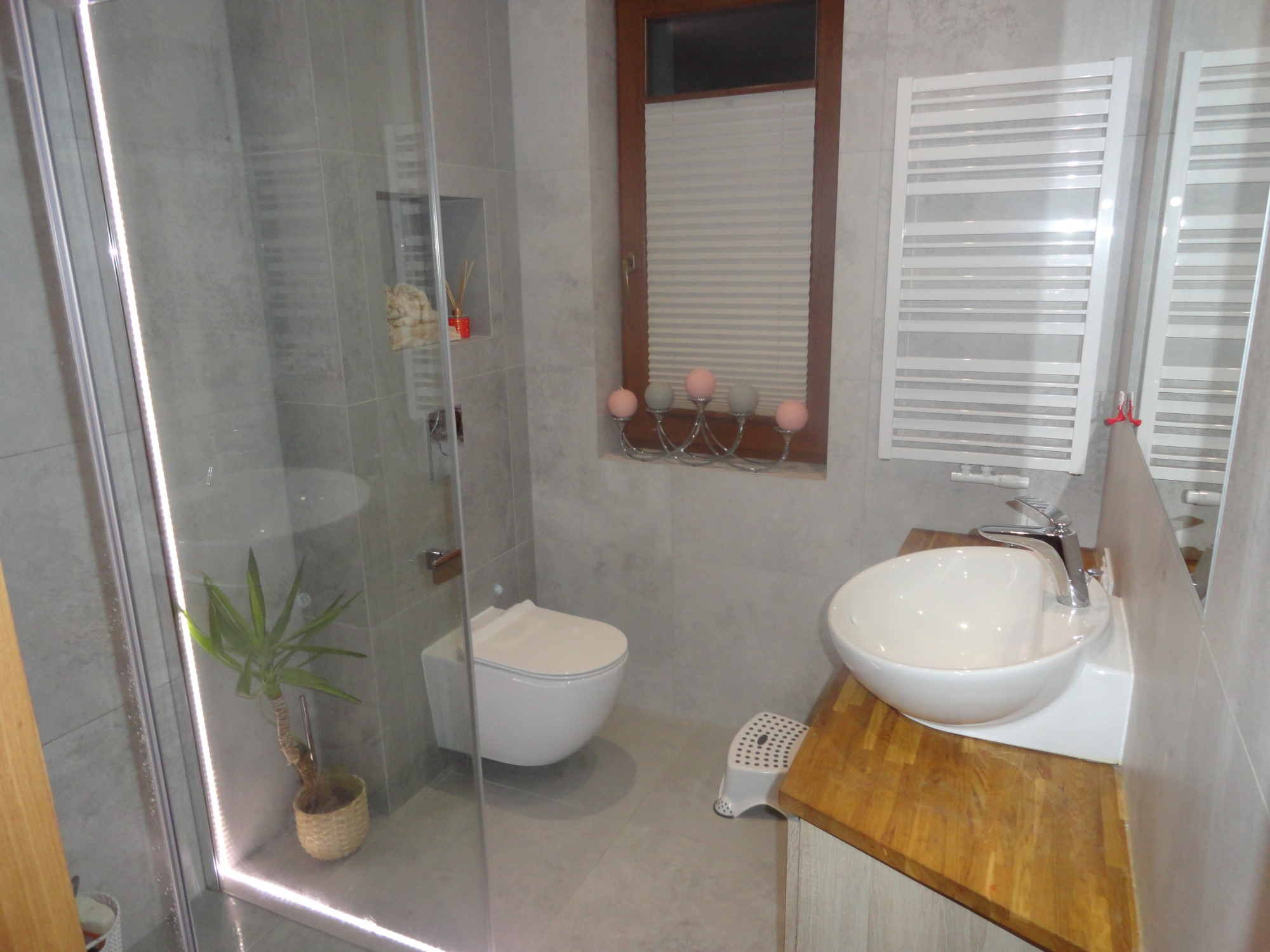 Nowoczesna łazienka z szarą glazurą, kabina prysznicowa z rośliną w rogu, podwieszana toaleta i umywalka nablatowa na drewnianym blacie, oświetlona listwą LED wzdłuż kabiny.