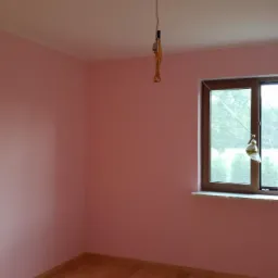 Pokój po malowaniu na różowo w Czerniejewie, widoczne drewniane okno z uchylonym skrzydłem, drewniana podłoga i wiszący kabel z żarówką.